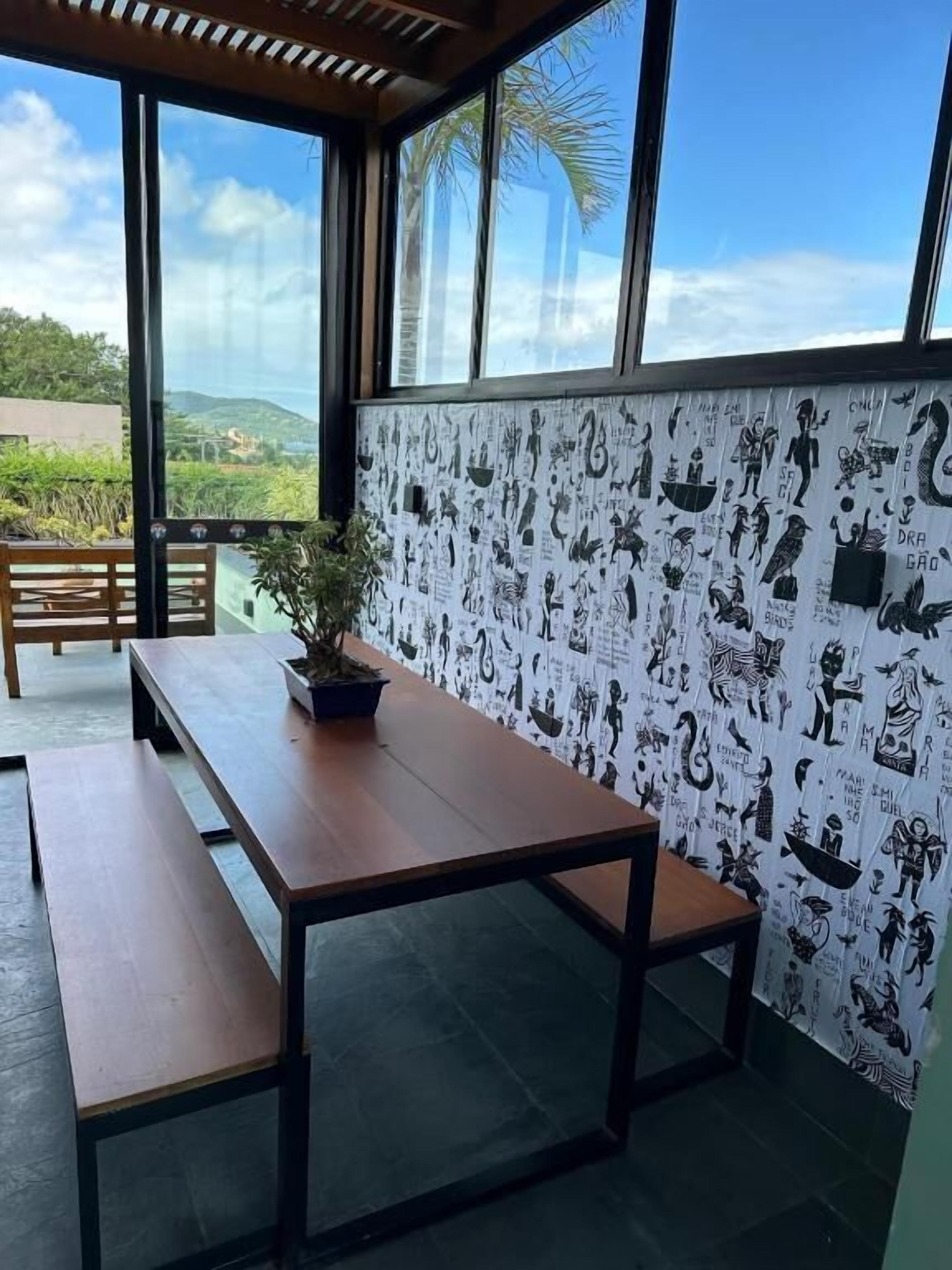 A room with a large window overlooking a scenic view, a wooden table with benches, and a wall decorated with black and white illustrations.