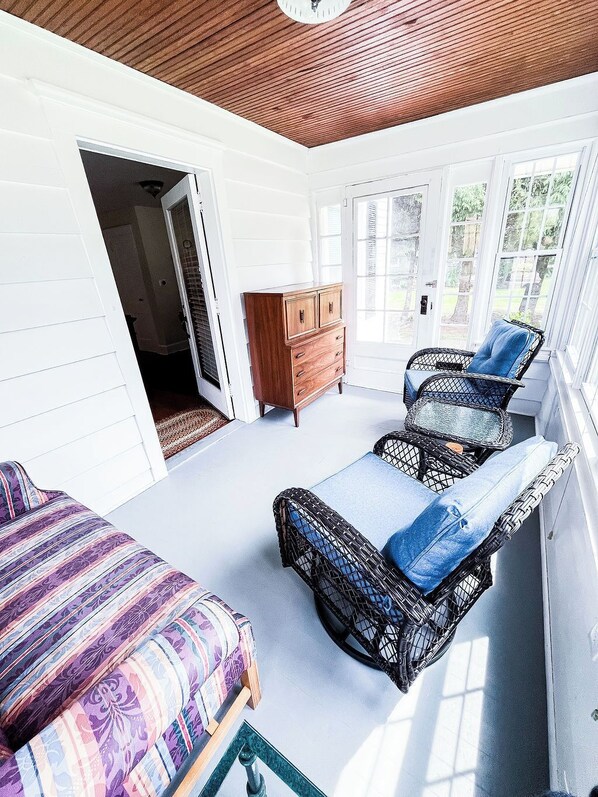 A room with a wooden ceiling, a bed with a patterned bedspread, a wicker chair with a blue cushion, and a wooden dresser.