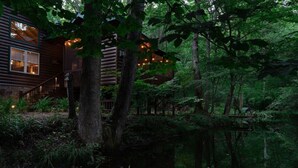 A rustic log cabin with a deck overlooking a pond, surrounded by dense forest. - Olivers Hideaway | on Fightingtown Creek (Cherry Log)