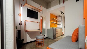 A compact living space with a bed, a small stool, a TV mounted on the wall, and a kitchenette in the background. - INDIE UNIVERSE  (Medellín)