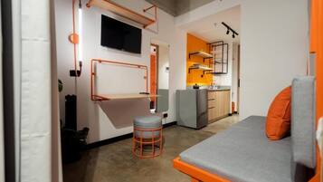 Un espacio habitable pequeño con cama, un taburete, televisor montado en la pared y una kitchenette al fondo.