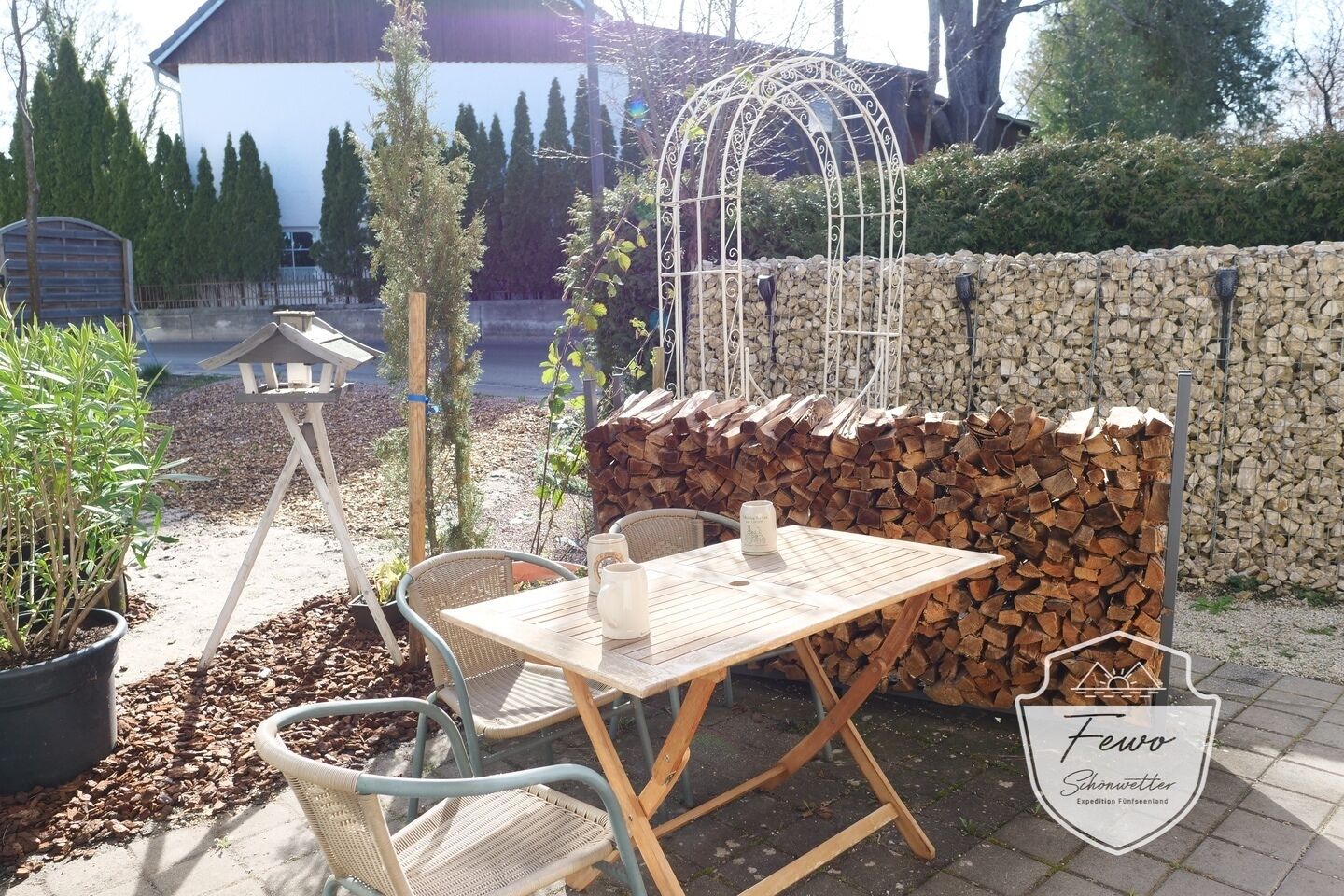 Une terrasse extérieure avec une table et des chaises en bois, une structure métallique décorative et une pile de bois de chauffage.