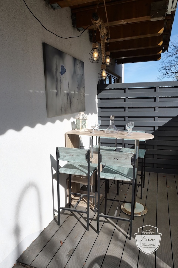 An outdoor patio with a wooden table and chairs, a hanging light fixture, and a wall-mounted painting.