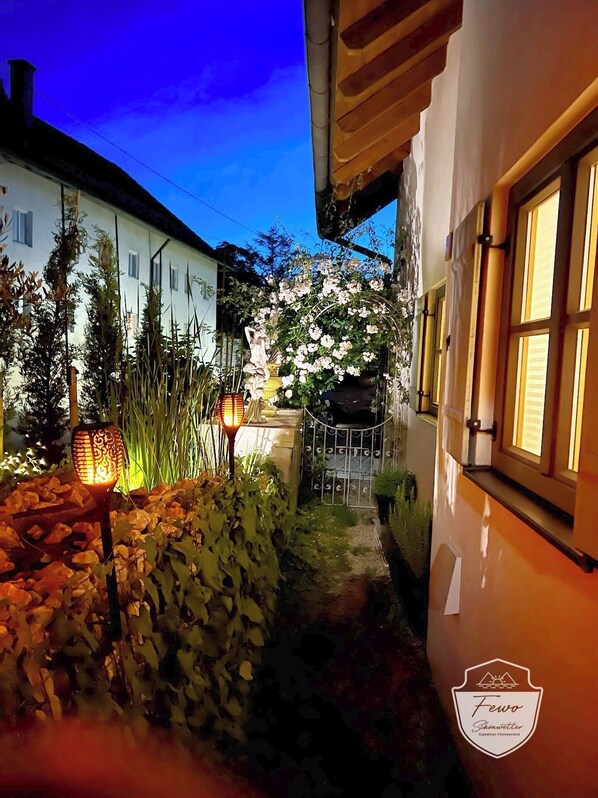 Un sendero estrecho que lleva a un edificio blanco con jardín, una lámpara decorativa y una reja.