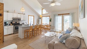 Une cuisine moderne avec des meubles blancs, un îlot central et des chaises en bois. On y trouve un coin repas avec une table et des chaises blanches, ainsi qu’un salon avec un canapé beige et une table basse blanche.