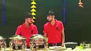 Um palco com duas pessoas de camisa vermelha, uma mesa com recipientes de comida e um fundo decorado com enfeites pendurados.