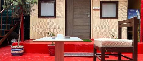 A red-tiled outdoor area with a table and a chair, a wooden door, and two framed windows.