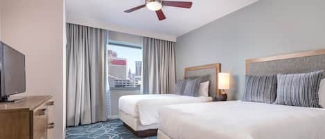 A hotel room with two beds, a ceiling fan, a TV, and a view of the city through the window.