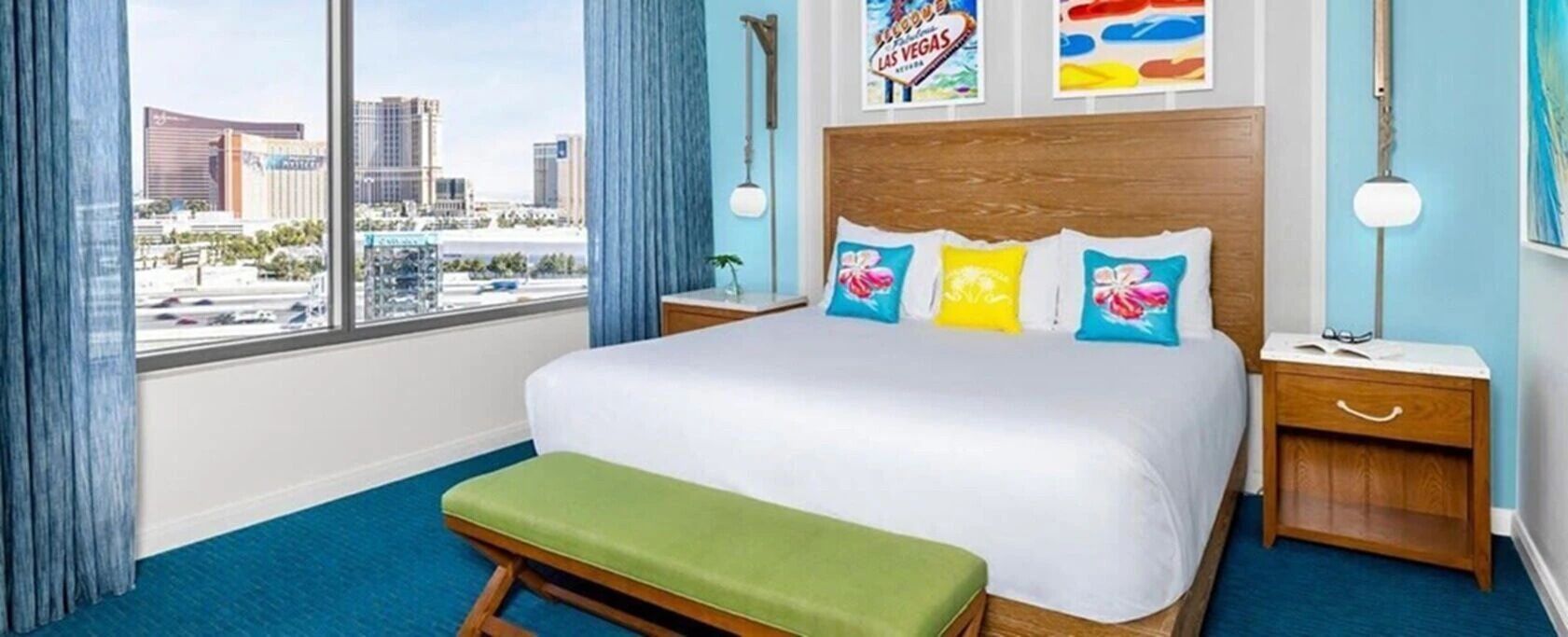 A hotel room with a large bed, a bench, a nightstand, a lamp, and a view of the cityscape.