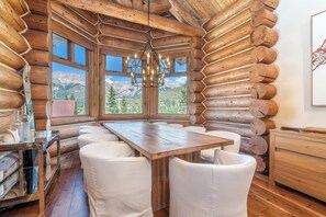 Interior de una cabaña rústica con una mesa de comedor de madera, sillas blancas acolchadas, una araña y ventanas amplias con vistas a las montañas.