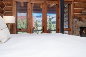 Une chambre avec un grand lit, des murs en bois, une cheminée en pierre et une vue sur la montagne par une porte vitrée.