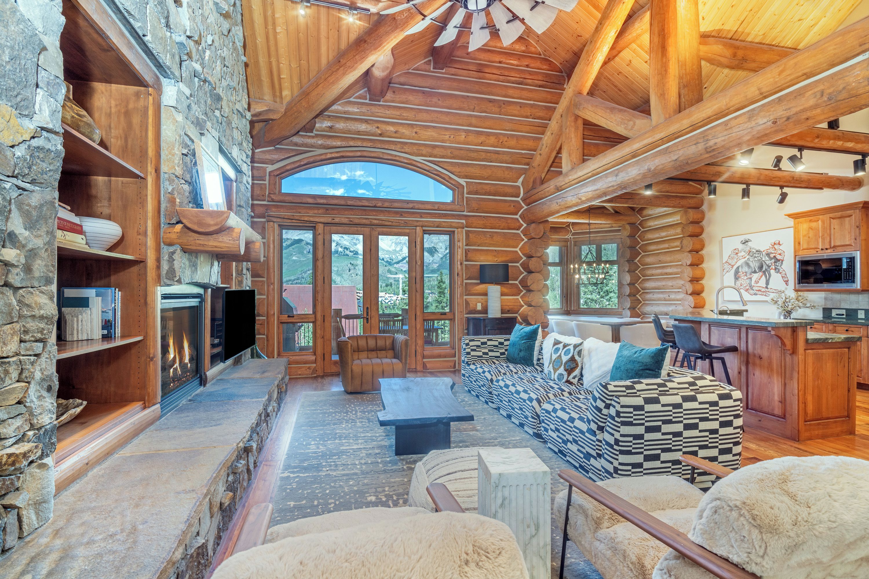 L’intérieur d’une cabane en rondins, avec une cheminée en pierre, une grande fenêtre donnant sur un paysage montagneux et un coin salon confortable agrémenté de coussins à motifs.