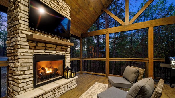 A stone fireplace with a flat-screen TV above it, surrounded by wooden deck furniture and a large window overlooking a forest. - NEW Listing Discounts! 1BR w/ loaded amenities! (Broken Bow)