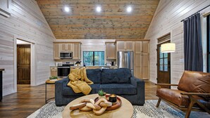 A spacious living area with a sofa, coffee table, and leather armchair. A kitchen with wooden cabinets and appliances is visible in the background. - NEW Listing Discounts! 1BR w/ loaded amenities! (Broken Bow)