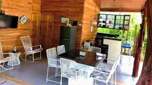 Une cabane en bois comprenant un coin repas avec des chaises en métal blanc et une table à plateau en verre, une cuisine avec une fenêtre et un téléviseur fixé au mur.