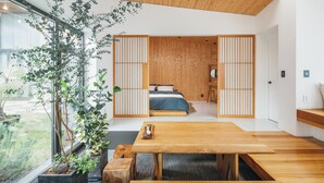A modern room with a wooden dining table, a bed with a blue bedspread, and a large window with a view of greenery.