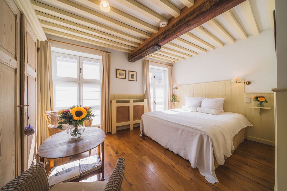 A bedroom with a wooden ceiling, a bed, a small table with a vase of flowers, and a window with curtains.
