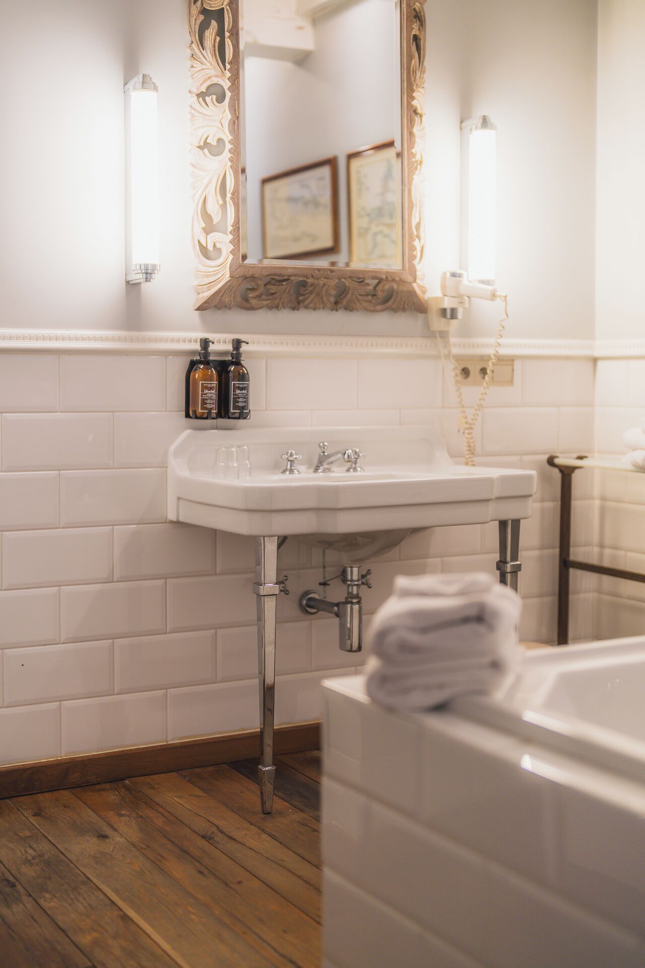 Un baño con pared de azulejos blancos, un lavabo blanco con dos grifos y suelo de madera.