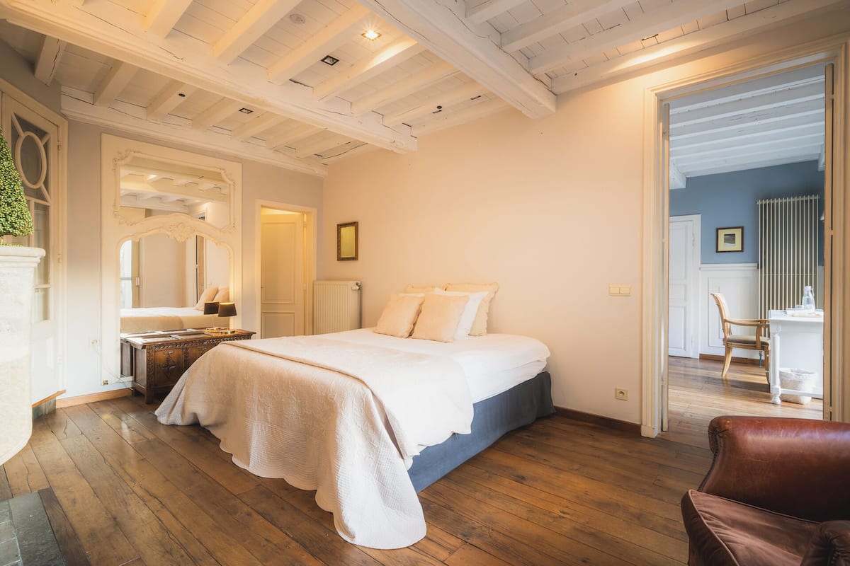 A bedroom with a bed, wooden flooring, a mirror, a dresser, and a chair.