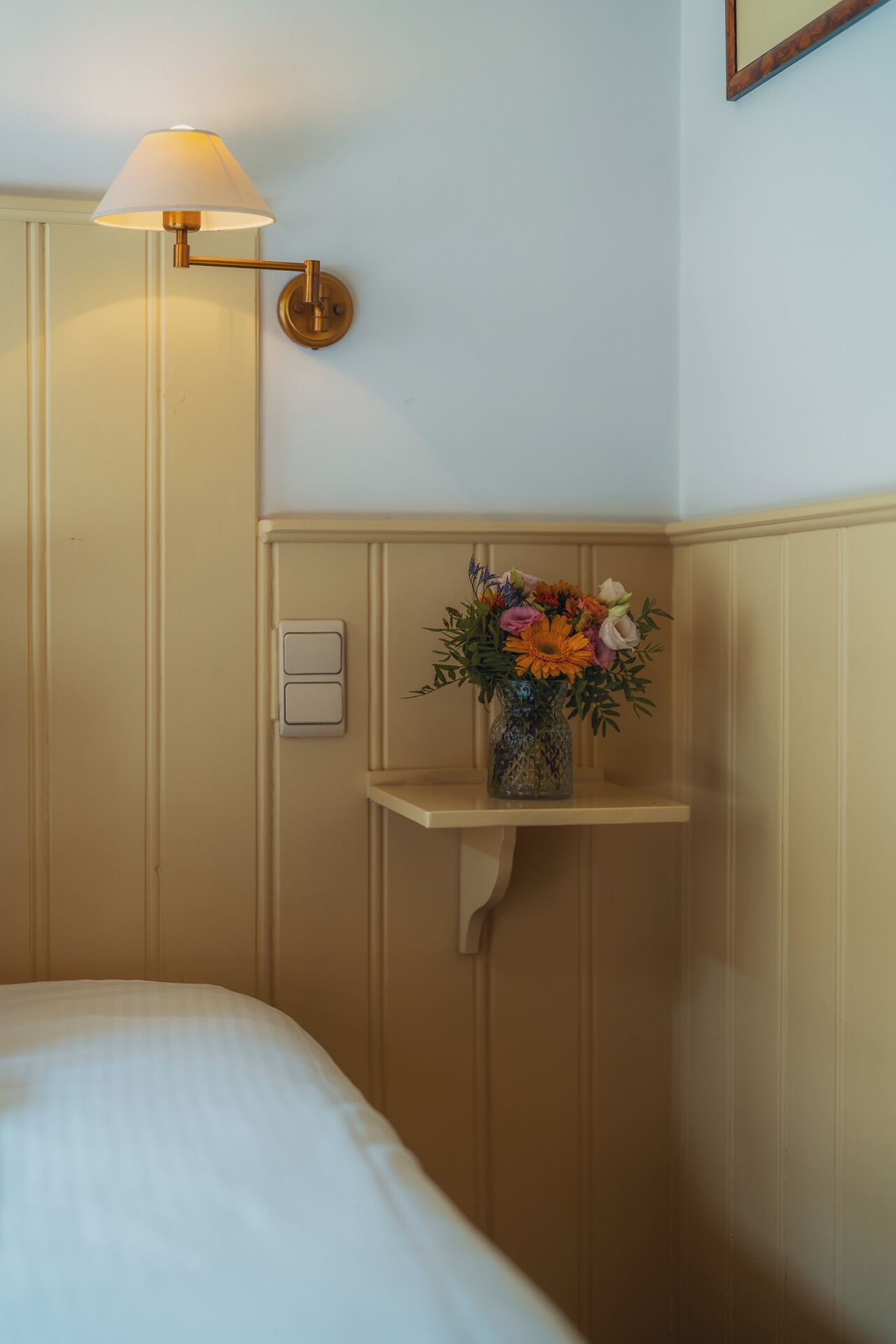 Une chambre d’hôtel avec un lit, un luminaire fixé au mur et une étagère agrémentée d’un arrangement floral.