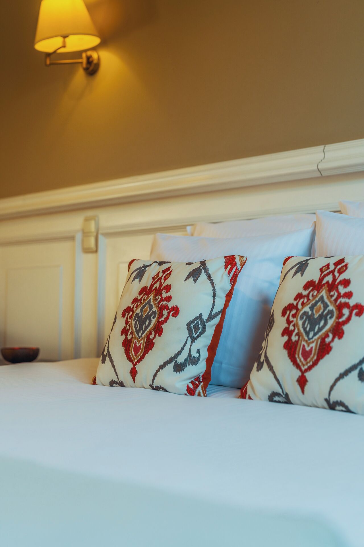 A bed with white bedding and patterned pillows, a wall-mounted lamp, and a white headboard.