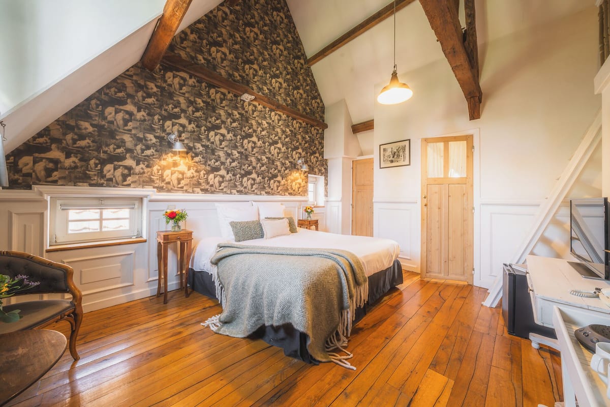 A cozy bedroom with a sloped ceiling, wooden beams, a large bed, a desk, and a window with a view.