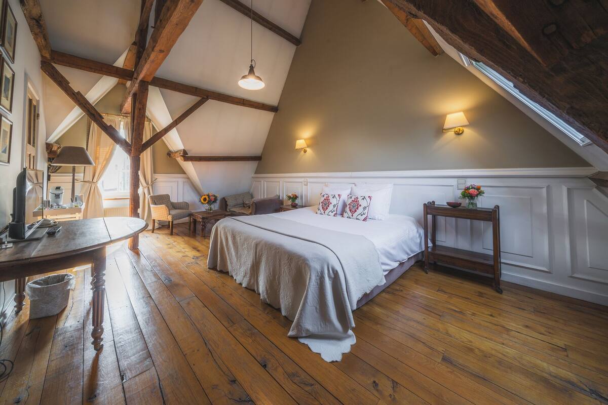 A bedroom with a sloped ceiling, wooden beams, a large bed with white bedding, a bedside table with a vase of flowers, a chair, and a desk with a lamp.