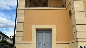 Un bâtiment à la façade beige, avec une porte double grise et un petit balcon au-dessus, ainsi qu'un sol recouvert de carreaux décoratifs.