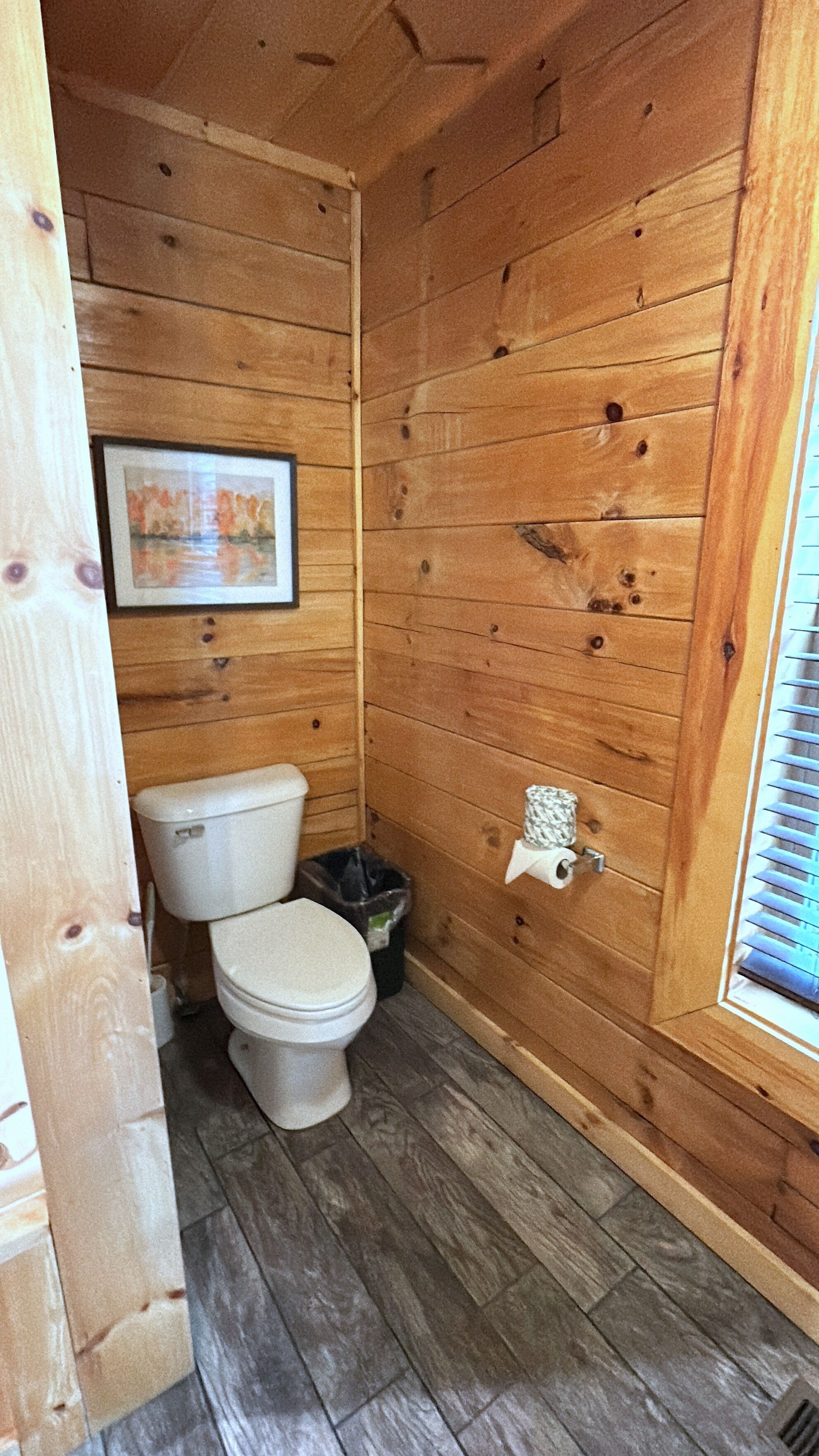 Ein kleines Badezimmer mit Holzvertäfelung, Toilette, Mülleimer und einem Fenster mit Jalousien.