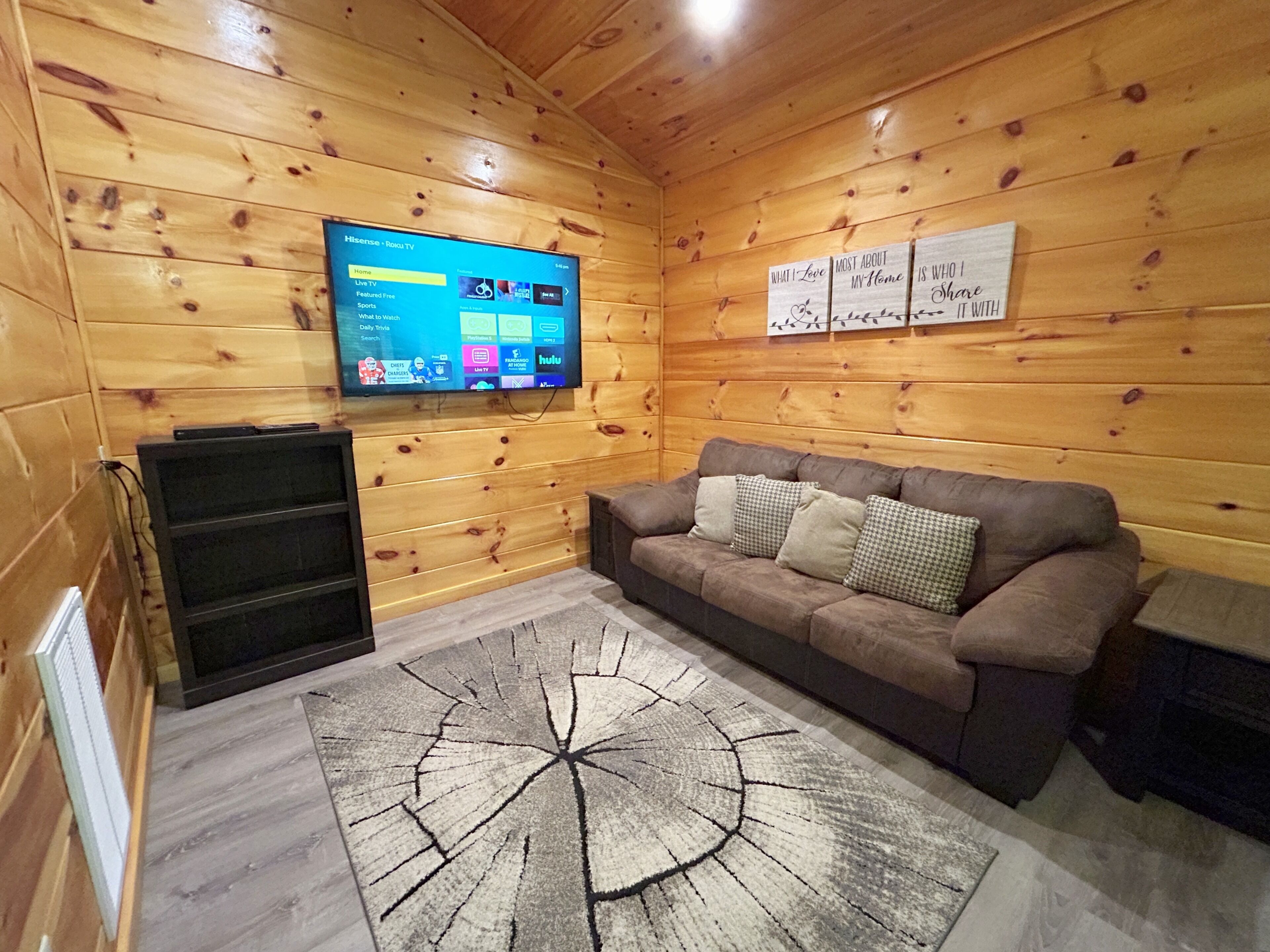 Ein gemütliches Zimmer mit Holzvertäfelung, einem an der Wand montierten Flachbildfernseher, einem braunen Sofa mit Kissen und einem einzigartigen, runden Teppich.