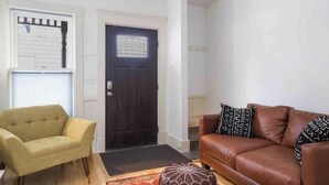 Un salon avec une porte en bois foncé, un fauteuil jaune, un canapé en cuir marron agrémenté de coussins à motifs, et un tapis rouge et orange.