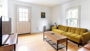 Een woonkamer met een houten vloer, een gele bank, een salontafel, een televisie en een deur met een raam.