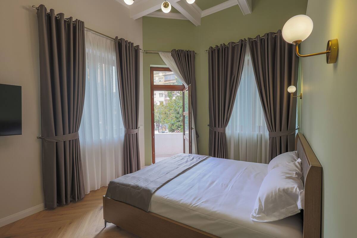 A bedroom with a bed, curtains, a wall-mounted light fixture, and a view through the window.