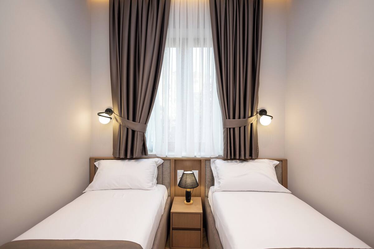 A hotel room with two single beds, a nightstand with a lamp, and curtains.