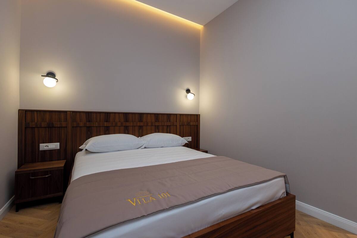 A neatly made bed with a brown bedspread, a wooden headboard, and two wall-mounted nightstands with light fixtures.
