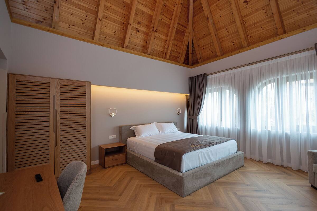 A bedroom with a wooden ceiling, a bed with white bedding, a nightstand, a desk, and a chair.
