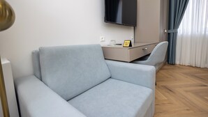 Una habitación de hotel moderna con un sofá de color gris claro, un televisor de pantalla plana montado en la pared, una mesita con un vaso y un cartel, y un suelo de madera con diseño de espiga.