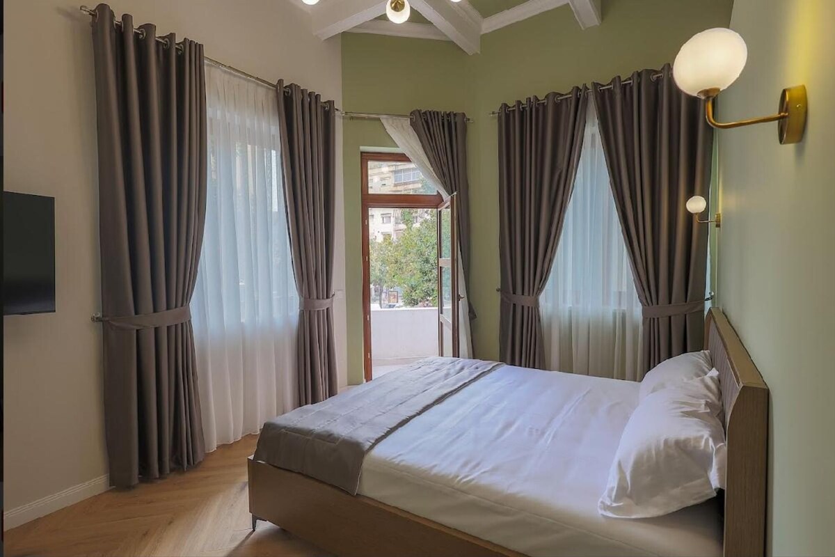 Un dormitorio con cama, cortinas, una lámpara fijada a la pared y vistas a través de una puerta abierta.