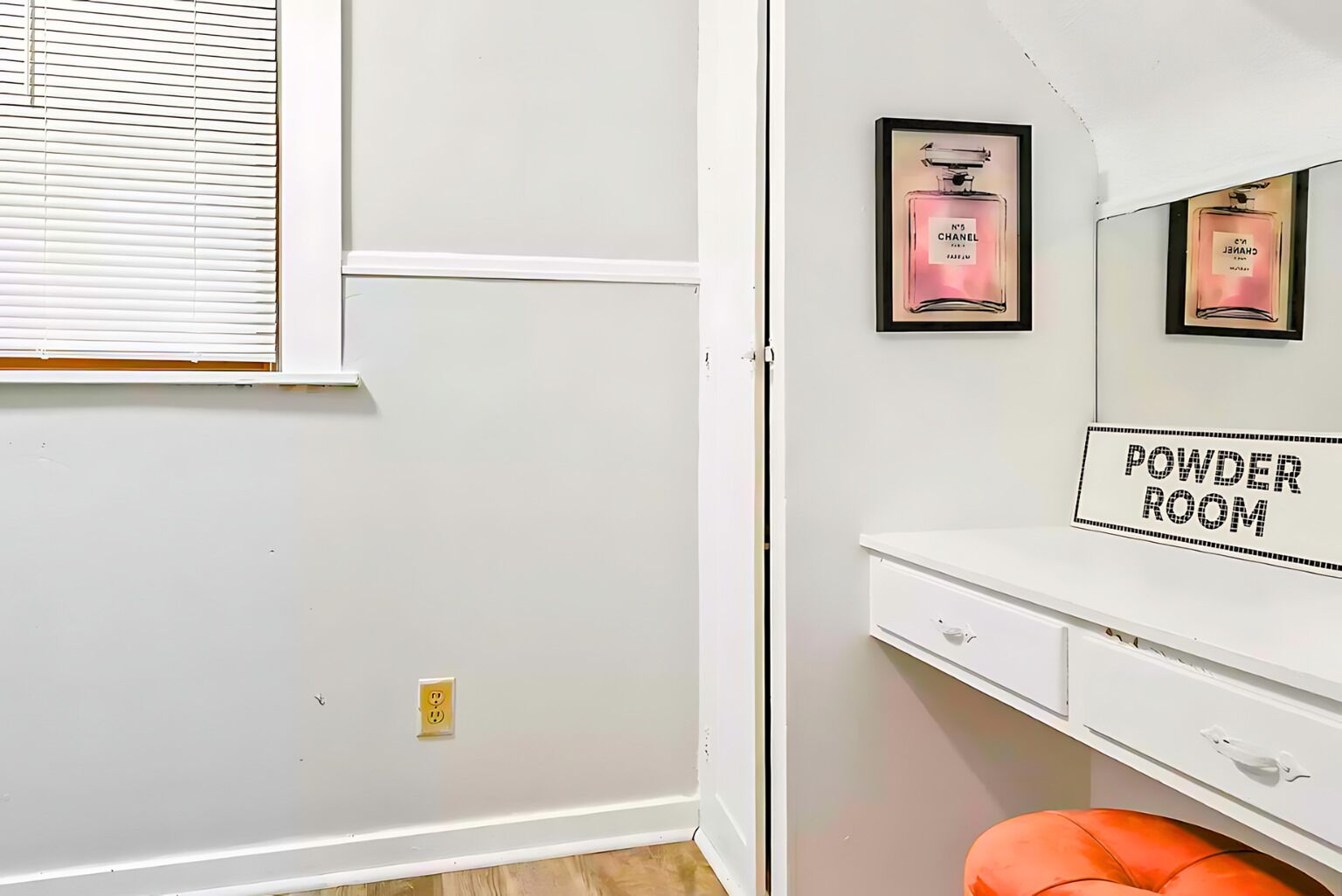 Una habitació blanca amb un cartell que diu 'Powder Room', una finestra amb persianes i dues ampolles de perfum emmarcades a la paret.