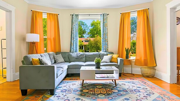 Ein Wohnzimmer mit grauem Sofa, einem Couchtisch und einem gemusterten Teppich. Zwei Fenster mit Vorhängen, eine Lampe und ein kleiner Tisch mit einer Pflanze sind ebenfalls vorhanden.