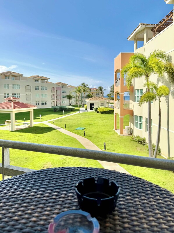 A balcony with a woven table, a small black object on it, and a view of a courtyard with buildings and palm trees.