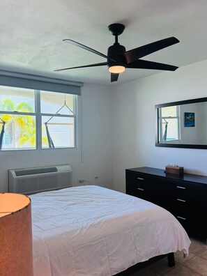 Een slaapkamer met een bed, plafondventilator, raam, verwarming, dressoir en spiegel.