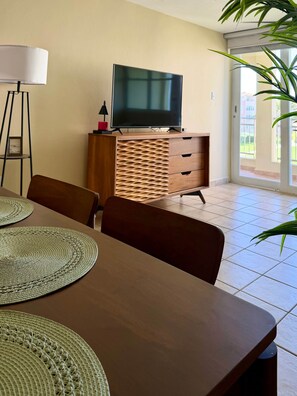 A dining area with a wooden table, woven placemats, a television on a wooden cabinet, and a plant. - #4 Beach Apt 2BR, 2BA - Jobos, Montones, Shacks (Isabela)