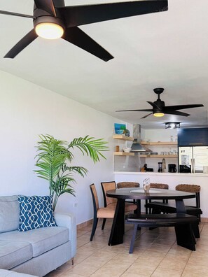 Een moderne woonkamer met een grijze bank, een eettafel met stoelen, een plafondventilator en een keuken met een koelkast en planken.