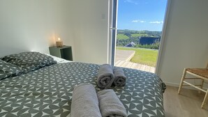 A bedroom with a large bed, a nightstand with a lamp, and a view of a green landscape through a glass door.