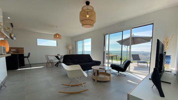 A modern living room with a black sofa, white armchairs, and a wooden rocking chair. There is a dining area with a table and chairs, and a kitchen area with a counter and appliances. Large windows provide a view of the outdoors.