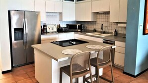 A modern kitchen with white cabinets, a stainless steel refrigerator, a black countertop, and two bar stools. - #17 REAL Beachfront 3BR Retreat @ Isabela Beach Ct (Isabela)