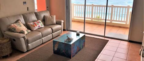 A living room with a sofa, a coffee table, and a view of the ocean through a sliding glass door.