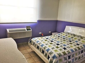 A bedroom with a bed, a bedside table, a window with blinds, and an air conditioning unit.