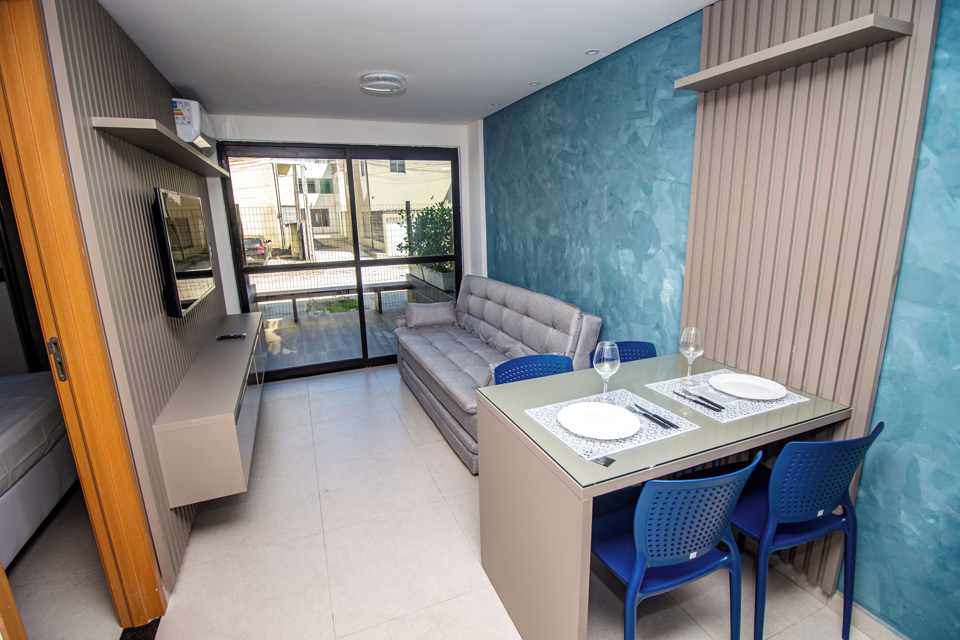 Una sala de estar moderna con un sofá, una mesa de comedor de vidrio con sillas azules y un televisor en un mueble empotrado.